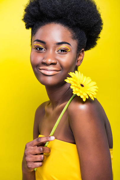 Smiling african american girl holding flower isolated on yellow - 写真・画像