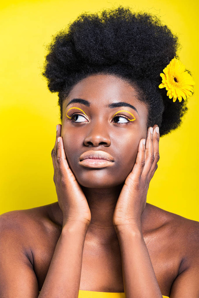 Dreamy african american woman with flower in hair looking away isolated on yellow - Foto, imagen