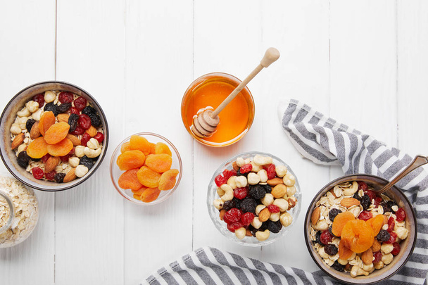 top view of bowls with cereal, dried berries and nuts served for breakfast with dried apricots and honey near striped cloth on white wooden table - Photo, Image