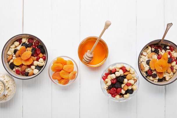 top view of bowls with cereal, dried apricots and berries, honey and nuts served for breakfast on white wooden table - Fotó, kép
