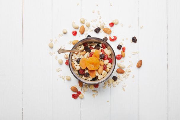 top view of bowl full of muesli with dried apricots, berries and scattered nuts around on white wooden table - Photo, Image