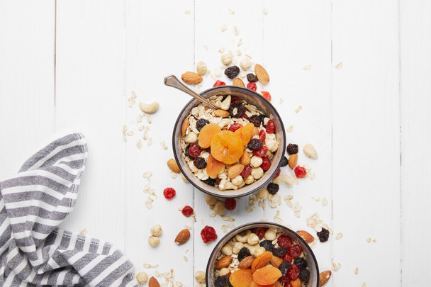 bovenaanzicht van kommen met muesli, gedroogde abrikozen en bessen en noten geserveerd voor het ontbijt met verspreide ingrediënten op witte houten tafel - Foto, afbeelding