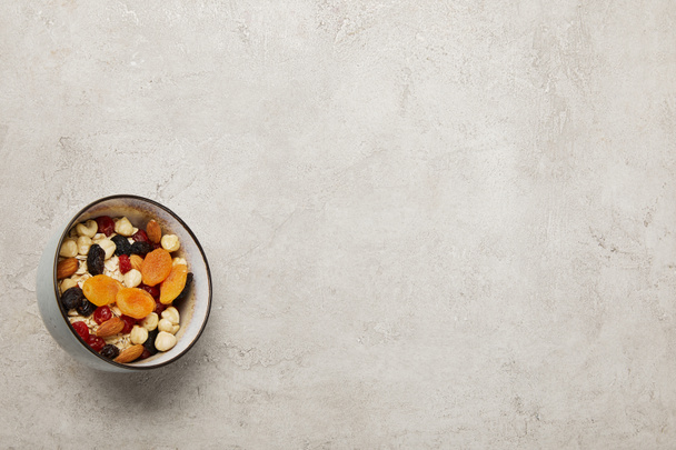 bovenaanzicht van schaal met muesli, gedroogde abrikozen en bessen, noten op getextureerde grijze ondergrond - Foto, afbeelding