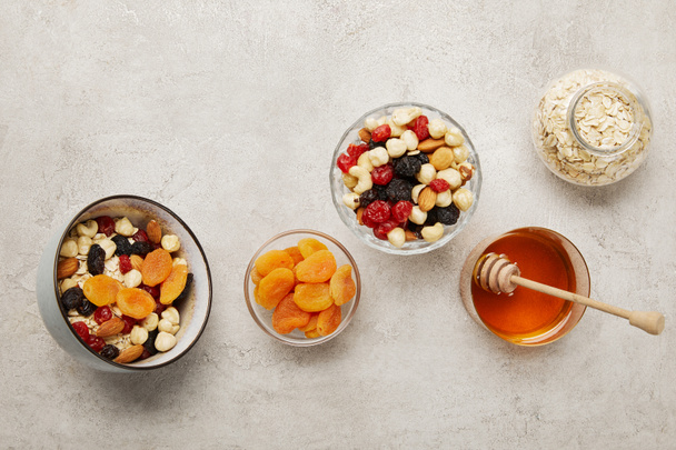 top view of bowls with muesli, dried apricots and berries, nuts and honey on textured grey surface - Photo, Image