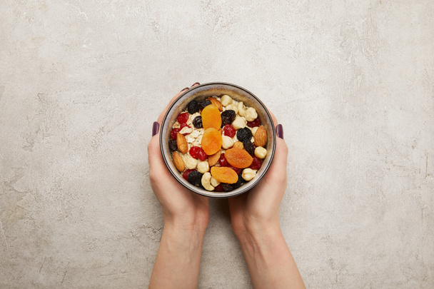 cropped view of woman holding bowl with muesli, dried apricots and berries, nuts on textured grey surface - Foto, imagen