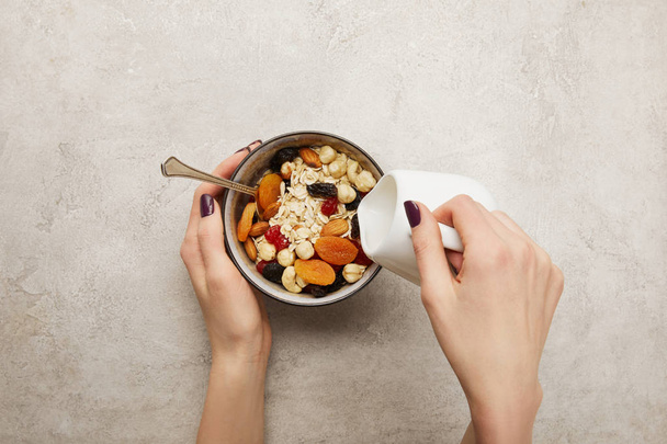 cropped view of woman pouring milk from milk jug into bowl with muesli, dried apricots and berries, nuts on textured grey surface - Photo, Image