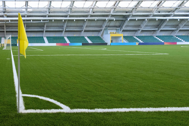 Corner of a soccer field in an indoor stadium. Yellow flag, white marking on green grass. Spectator stands in the background. Copy space, mockup for billboards. - Photo, Image