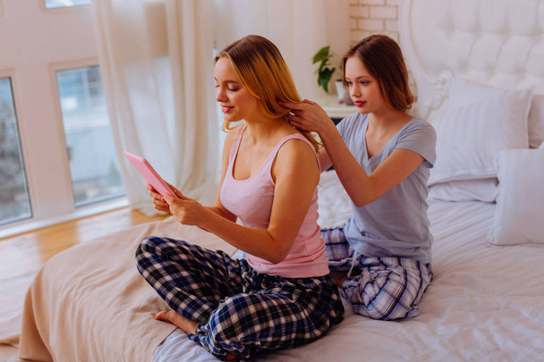 Younger sister doing hairstyle for her busy older sister using tablet - Photo, Image