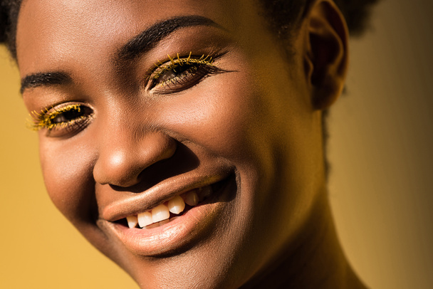 Hymyilevä afrikkalainen amerikkalainen nainen, jolla on keltaiset silmäripset, katsoo kameraa ruskealla
 - Valokuva, kuva