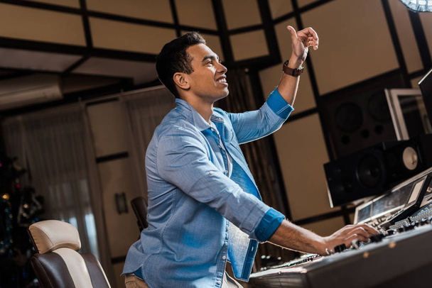 lächelnder Mixed Race Soundproduzent gestikuliert während er im Tonstudio arbeitet - Foto, Bild