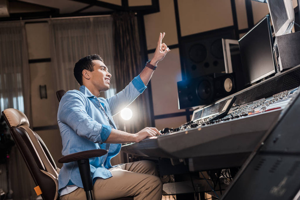 schöner Mixed-Race-Soundproduzent zeigt Siegeszeichen, während er im Tonstudio arbeitet - Foto, Bild