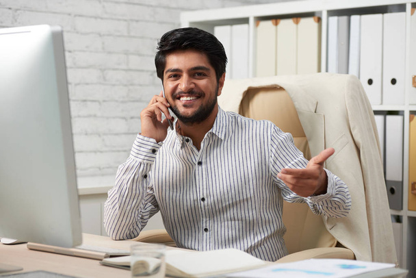 Onnellinen nuori komea liikemies puhuu puhelimessa ja toivottaa asiakkaat tervetulleiksi käymään toimistossaan
 - Valokuva, kuva