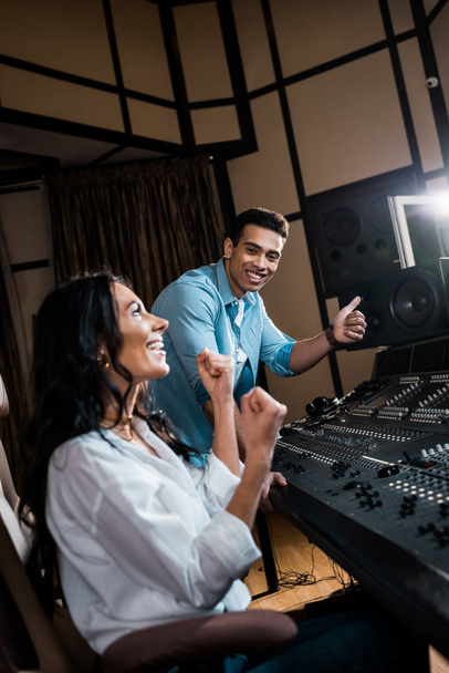 δύο χαρούμενοι πολυπολιτισμικοί παραγωγοί που δείχνουν χειρονομίες επιτυχίας στο στούντιο ηχογράφησης - Φωτογραφία, εικόνα