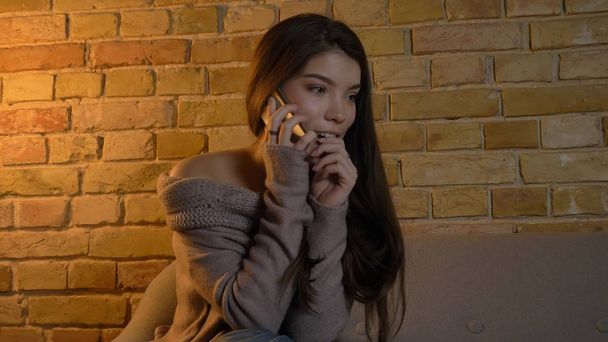Closeup shoot of young pretty caucasian female calling on the phone with excited facial expression while resting on the couch in a cozy apartment indoors - 写真・画像
