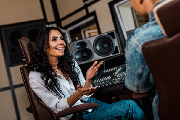 alegre productor de sonido atractivo hablando con colega de raza mixta en el estudio de grabación
 - Foto, Imagen