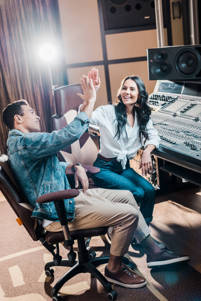 two cheerful multicultural sound producers giving five in recording studio while sitting near mixing console - Photo, Image