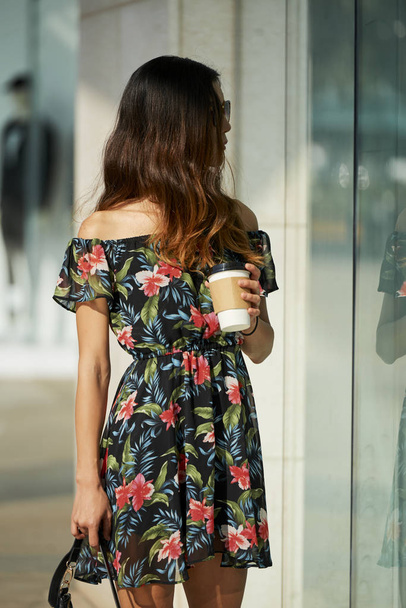 Elegantní žena v docela květinové šaty držící šálek horké nápoje a při pohledu na výlohy na ulici - Fotografie, Obrázek