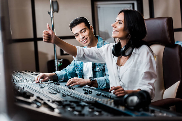 selective focus of cheerful sound produce showing thumb up while working at mixing console near smiling mixed race friend - Photo, Image
