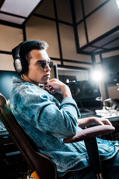 handsome serious mixed race sound producer in sunglasses and headphones sitting in recording studio - Foto, Imagem