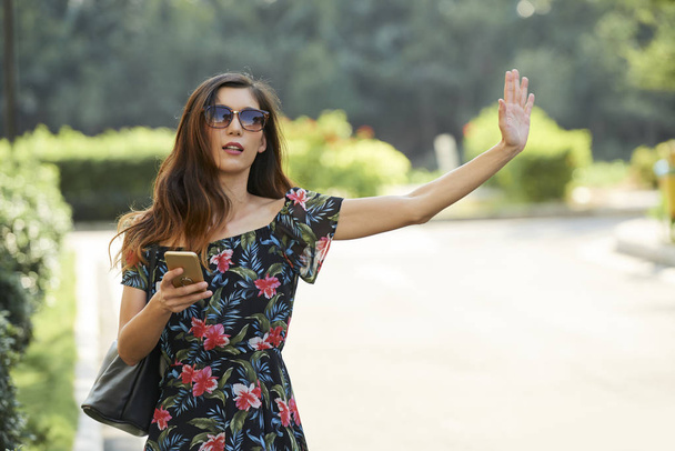 Encantadora mujer asiática en traje de moda sosteniendo el teléfono inteligente y saludando la mano mientras intenta detener el taxi cerca del parque
 - Foto, imagen