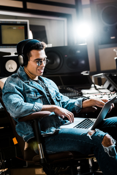 smiling mixed race sound producer in headphones using laptop in recording studio - Photo, Image