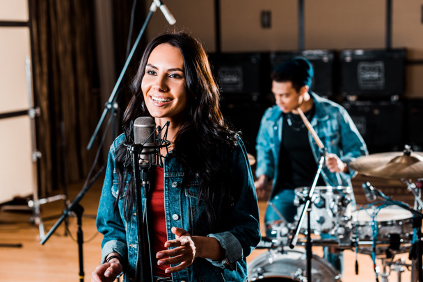 pretty woman singing while mixed race musician playing drums in recording studio - Photo, Image