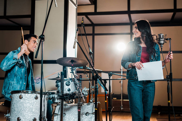 handsome mixed race musician at drums and attractive singer near microphone in recording studio - Foto, Imagem