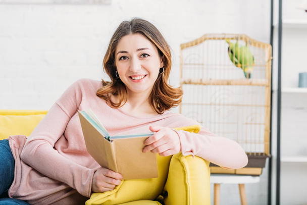 attractive woman holding book and looking at camera at home - Foto, imagen