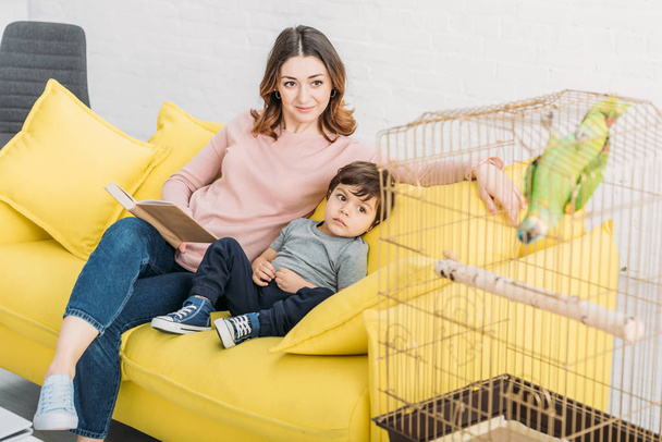 smiling mother with adorable son sitting on sofa near bird age with green parrot - Foto, Imagem