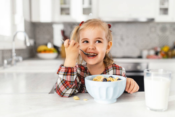 Hermosa niña desayunando con cereales, leche y arándanos en la cocina
 - Foto, imagen