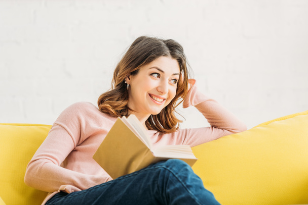 smiling attractive woman with book resting on yellow sofa at home - Foto, immagini