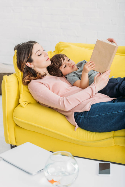 junge Mutter mit entzückendem Sohn liest Buch, während sie zusammen auf dem Sofa liegt - Foto, Bild
