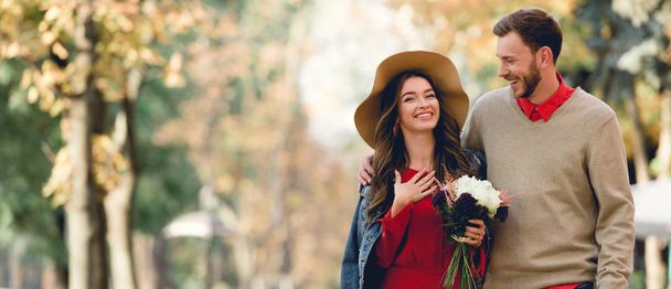 panoramatický záběr šťastného muže, který se při pohledu na přítelkyni usmívá a drží květiny  - Fotografie, Obrázek