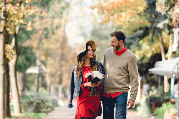 happy woman in hat smiling and holding flowers while looking at boyfriend  - 写真・画像
