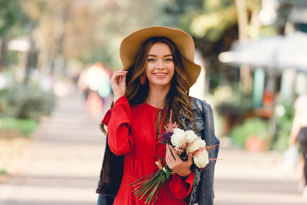 happy woman in hat smiling while holding flowers and looking at camera - Foto, Bild