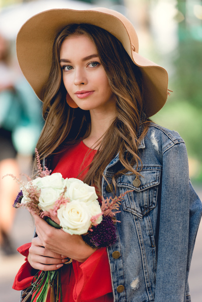 attractive girl in hat holding flowers and looking at camera - Fotó, kép