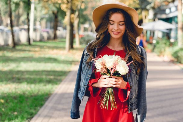 menina atraente no chapéu olhando para as flores, enquanto em pé no parque
  - Foto, Imagem