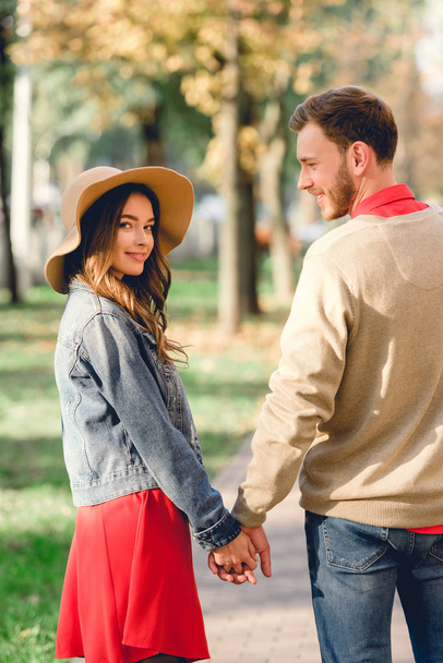 cheerful man looking at attractive girlfriend in hat while holding hands in park  - Fotografie, Obrázek