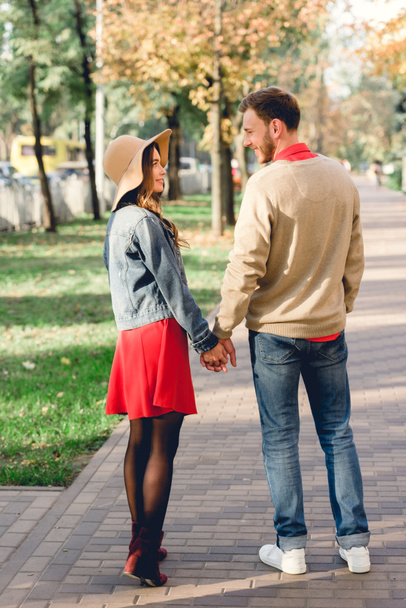 handsome man looking at happy girlfriend in hat while holding hands in park  - Valokuva, kuva