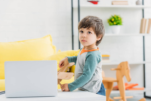 прелестный серьезный мальчик, стоящий рядом со столом с ноутбуком дома
 - Фото, изображение
