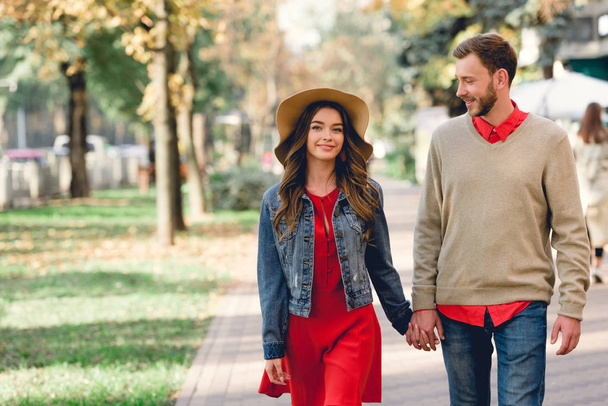 cheerful boyfriend looking at happy girlfriend in hat while holding hands in park  - Foto, imagen