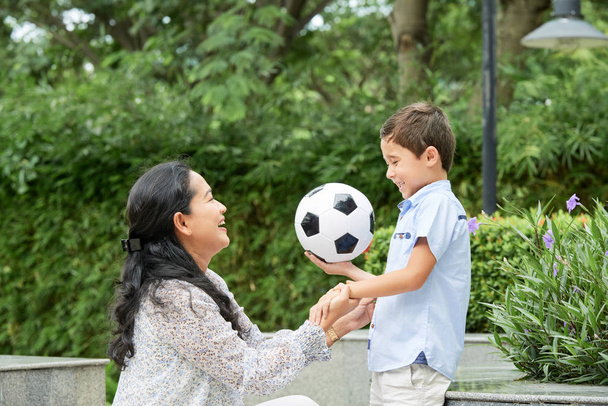 Vista lateral da mulher asiática alegre que apoia o menino bonito com bola de futebol ao passar o tempo no parque bonito junto
 - Foto, Imagem