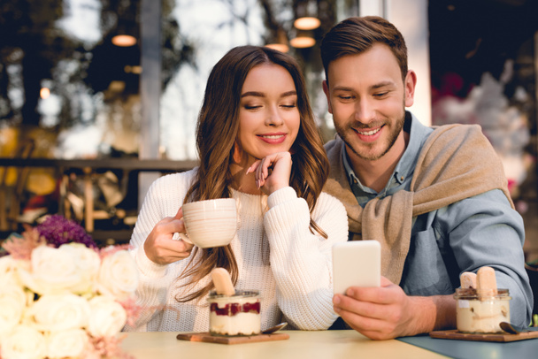 cheerful man looking at smartphone near attractive girl holding cup of coffee  - Photo, Image