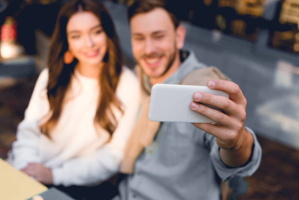 selective focus of smartphone in hand of cheerful man taking selfie with young woman  - Foto, Bild