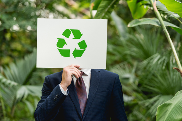 businessman holding white card with green recycling sign in front of face in greenhouse - Photo, image