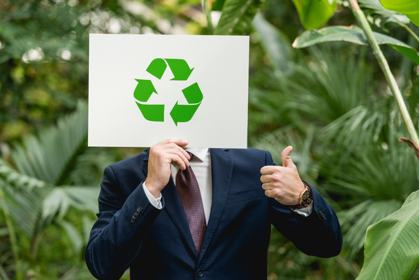 businessman holding white card with green recycling sign in front of face and showing thumb up in greenhouse - Photo, Image