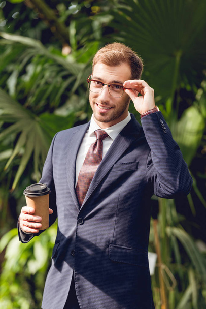 χαμογελαστός επιχειρηματίας σε κουστούμι, γραβάτα και γυαλιά κρατώντας καφέ για να πάει σε πορτοκαλύλλα - Φωτογραφία, εικόνα