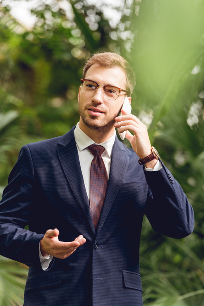 handsome businessman in suit, tie and glasses talking on smartphone in greenhouse - Фото, изображение