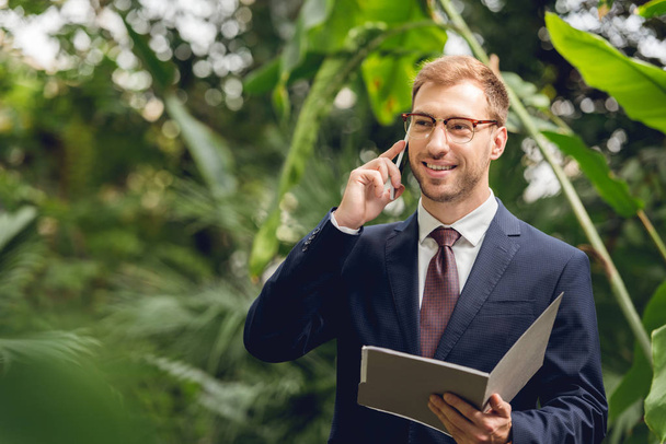 happy businessman in suit and glasses talking on smartphone and holding folder in greenhouse  - Foto, Imagem