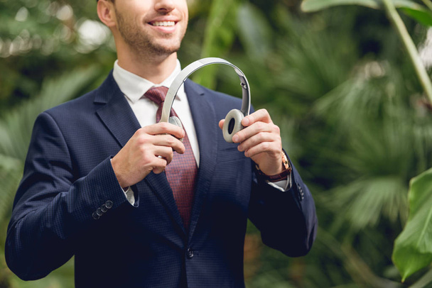 cropped view of smiling businessman in suit and tie holding wireless headphones in greenhouse - Fotoğraf, Görsel